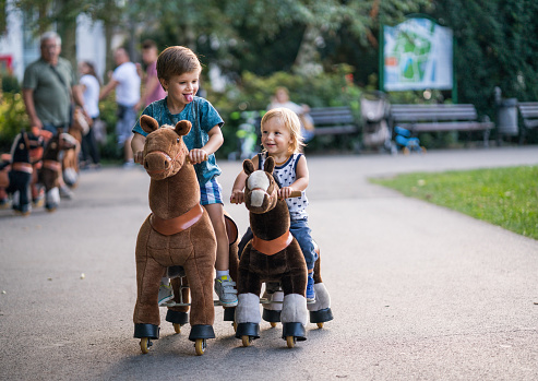 Two playful boys having fun in the park while riding toy horses. One of them is sticking out a tongue.