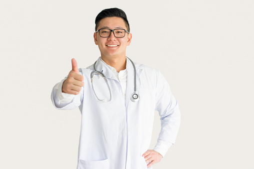 Smiling Asian doctor showing thumb up at camera. Young medical resident wearing glasses with ok gesture. Medicine, health care and approving concept