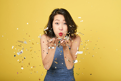 Cheerful young Asian woman blowing confetti into camera