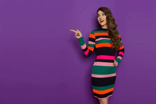 Beautiful young woman in colorful vibrant striped mini dress with long sleeves is looking away, pointing and talking. Three quarter length studio shot on purple background.