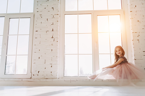 Cute little girl in beautiful dress is sitting on a window sill at light sunny room