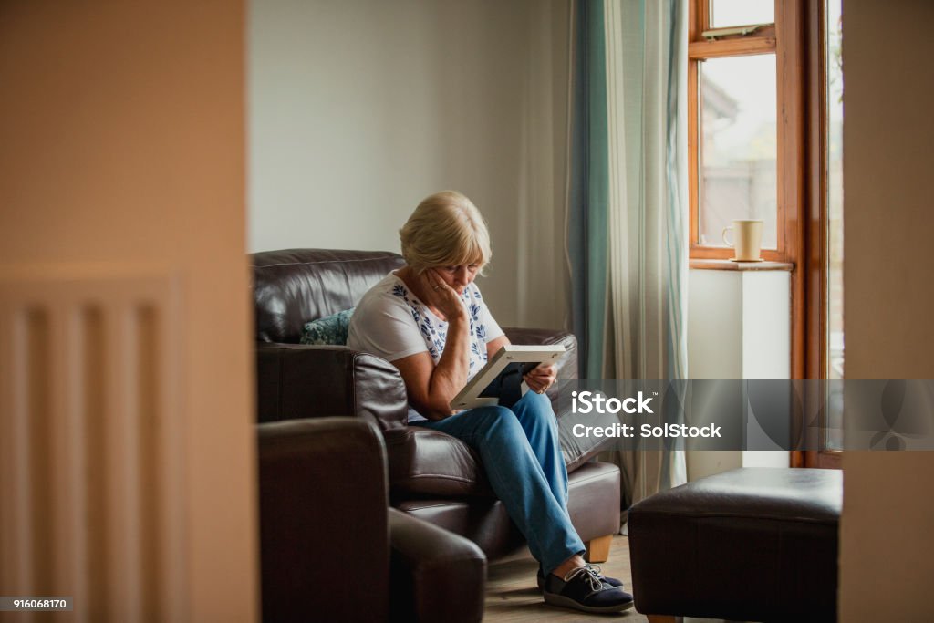 Senior Woman Looking at Photo Frame A senior woman sits in the living room and looks at a photo frame. Death Stock Photo