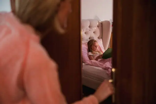 A woman checks on her daughter and finds her reading a book in her bedroom.