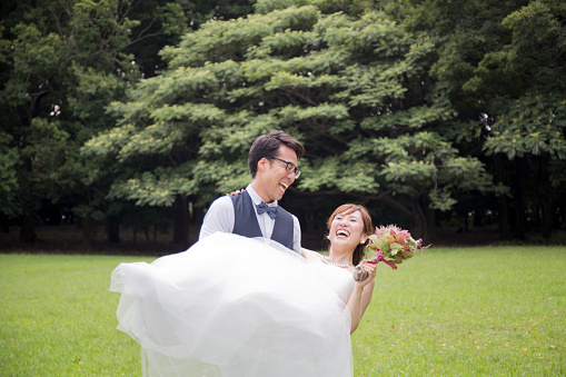 Japanese man carrying Japanese woman who is wearing wedding dress and holding bouquet in her hand.