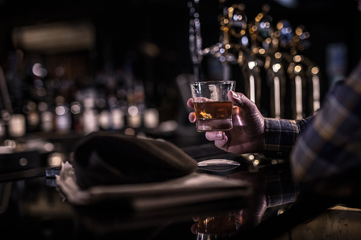 an hold man's hand holding a half filled whiskey glass, at a dark moody bar counter in a shallow depth of field