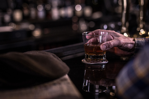 an old man hand holding a glass of whiskey at a dark seedy bar counter, with shallow depth of field