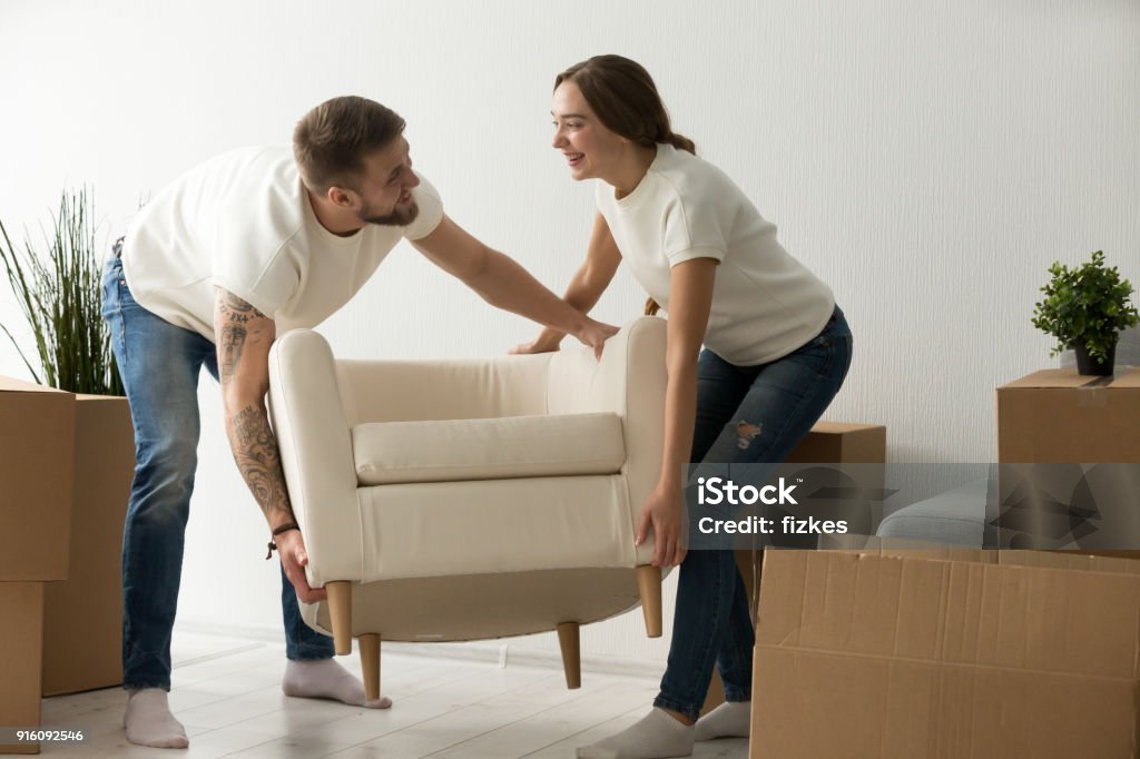 Young couple carrying chair together, placing furniture in new h Young couple carrying chair together, house improvement, modern furniture in new home concept, man and woman moving into own flat after relocation furnishing living room, remodeling and renovation Furniture Stock Photo