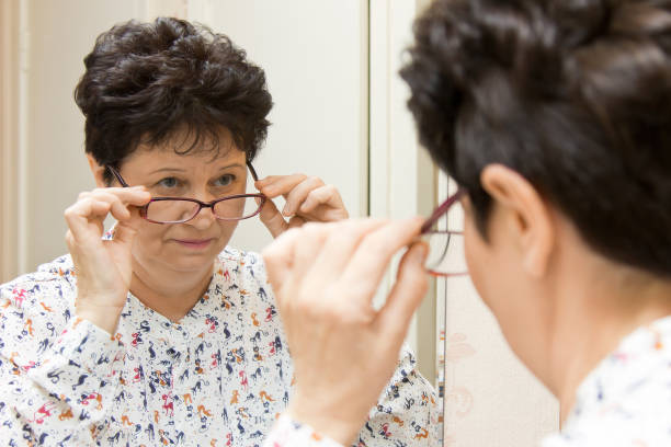 Senior woman trying on new eyeglasses and looking over the glasses in the mirror Senior woman trying on new eyeglasses and looking over the glasses at herself in the mirror person holding frame over face stock pictures, royalty-free photos & images