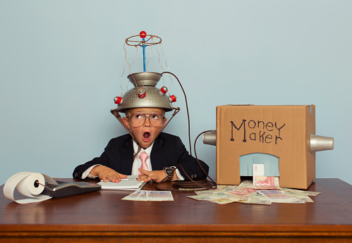 A young British boy dressed in business attire wears a homemade mind reading helmet attached to a money making machine. The money machine is making real British Pound Sterling from his head full of ideas. He is surprised at this.