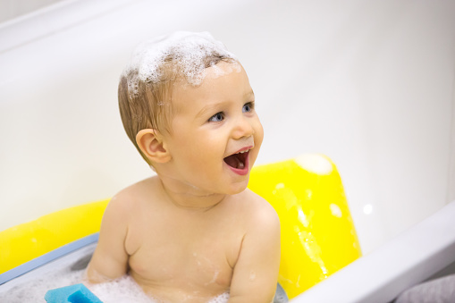 Excited child taking bath with toys and foam on head, surprised funny kid having fun in bathroom playing and laughing, cute happy toddler enjoying bath-time splashing in bathtub, baby bathing concept