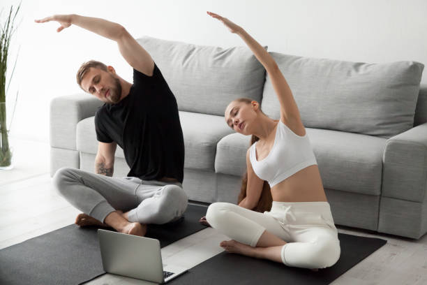 Sporty couple stretching watching fitness video tutorial online Sporty young couple warming up stretching watching fitness video tutorial online on laptop, fit man and woman doing workout at home sitting on mat in living room practicing yoga side bending exercise two men same clothes stock pictures, royalty-free photos & images
