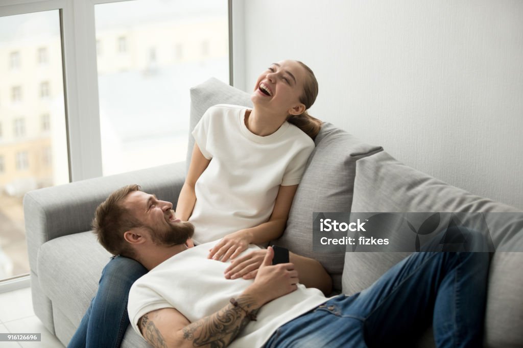 Cheerful happy couple laughing with joy relaxing at home togethe Cheerful happy couple laughing with joy relaxing at home on sofa together, positive man and woman having fun enjoying funny talk, boyfriend telling hilarious joke chatting with girlfriend on couch Adult Stock Photo
