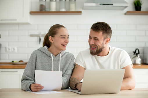 Young smiling happy couple talking at home in the kitchen with laptop papers, cheerful man and woman holding documents excited by planning new purchase, having fun enjoying positive news in letter
