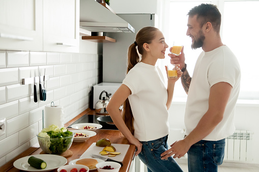 Young smiling couple preparing breakfast in cozy kitchen, loving man and woman drinking fresh orange juice or smoothie enjoying pleasant morning cooking together, healthy meal and lifestyle concept