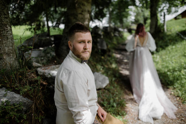 istock handsome young groom looking at camera and bride walking on mountain path in Alps 916132174