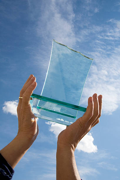 Glass award held up high by two hands in front of blue sky stock photo