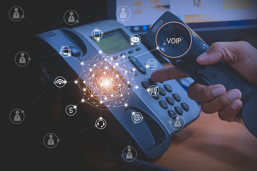 A close-up of a VOIP phone with a hand gripping the receiver, surrounded by digital icons representing communication functions like messaging, internet connections, and financial transactions.