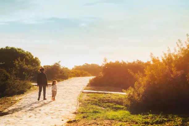 mother and daughter walk holding hands at sunset on a cobbled path and surrounded by greenery that leads to the beach