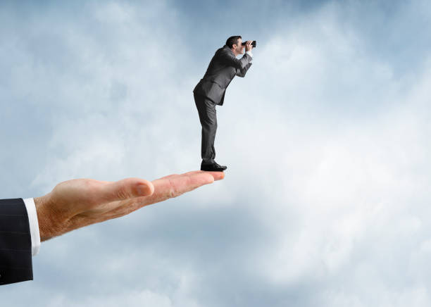 istock Businessman Looks Through Binoculars While Standing On Large Male Hand 916219698