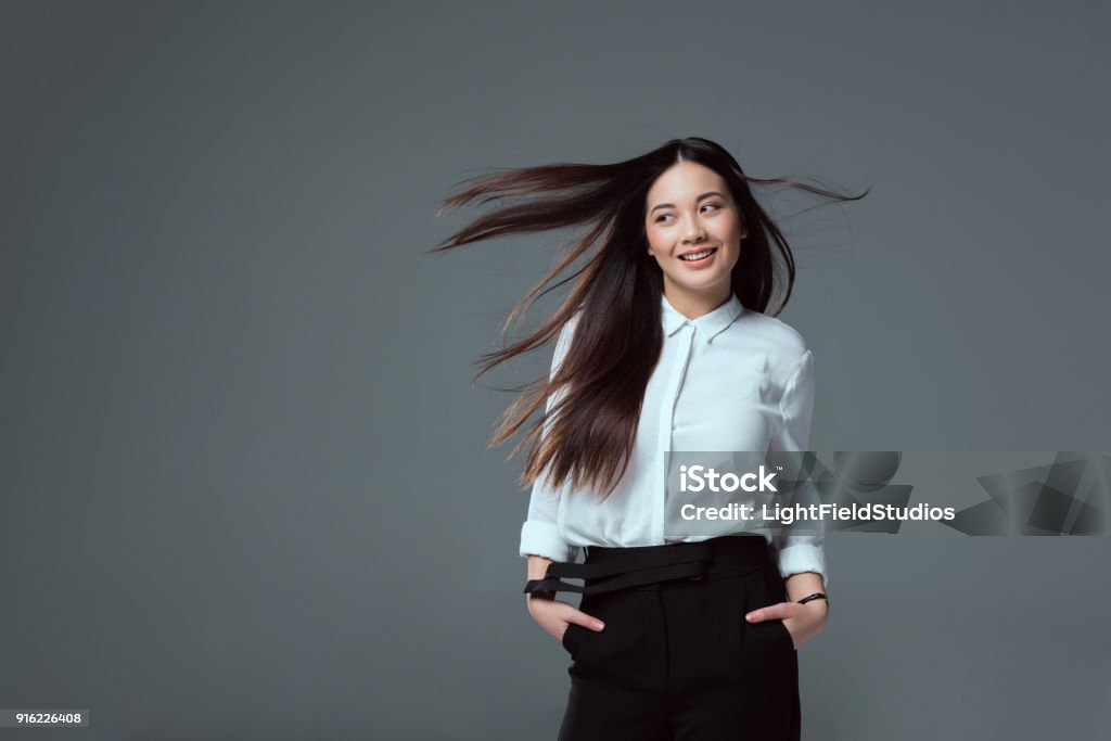 asian girl standing with hands in pockets and looking away isolated on grey beautiful smiling asian girl standing with hands in pockets and looking away isolated on grey Adult Stock Photo
