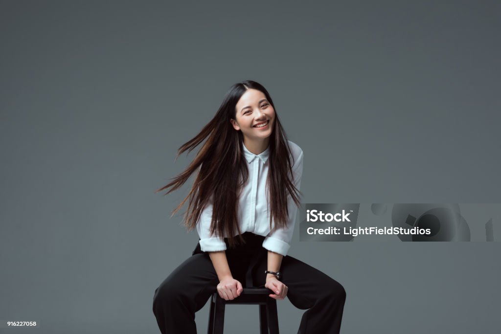 menina asiática atraente, sentado na cadeira e sorrindo para a câmera isolada em cinza - Foto de stock de Ásia royalty-free