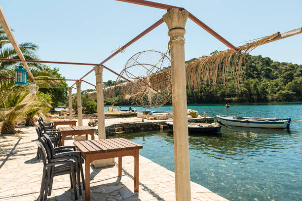 Empty chairs and tables at the water edge. Some small boats anchored at the stony pier.