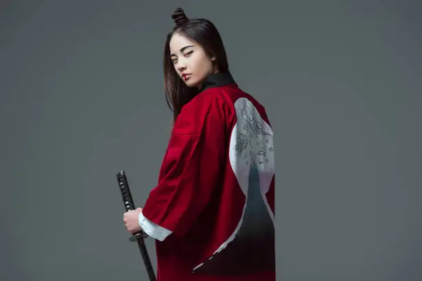 young woman in traditional japanese kimono holding katana and looking at camera isolated on grey