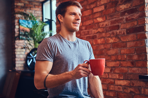 A stylish tattoed attractive man dressed in jeans and a t-shirt, sitting on a wooden chair, holding a cup of coffee. Looking away.