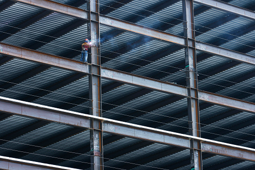 A welder welds on a steel i beam on a high rise building under construction.
