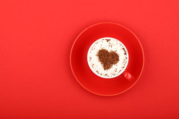 Close up one latte cappuccino coffee with heart shaped chocolate milk topping in red cup with saucer over background of red paper, elevated top view, directly above