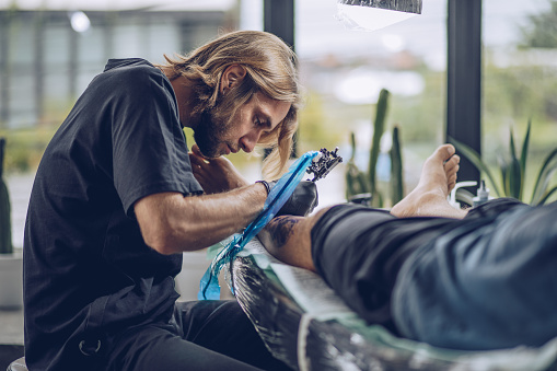 Two men in tattoo studio, man is tattooing a men's leg.