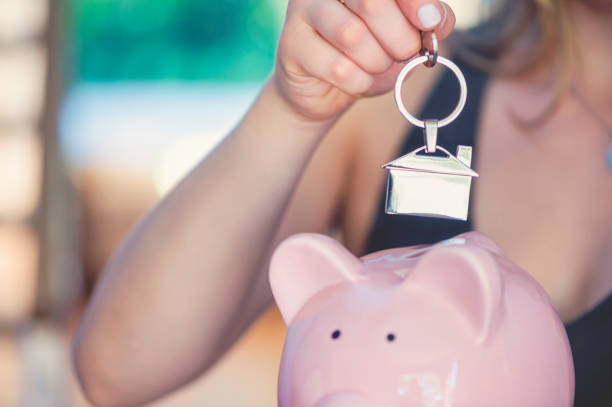 Woman holding house keys and a piggy bank. Woman holding house keys and a piggy bank. The key ring is house shaped. She is inserting or removing the keys from the piggybank. There is a home interior in the background. She is smiling and happy. Home ownership and savings concept. Copy space. australia house home interior housing development stock pictures, royalty-free photos & images