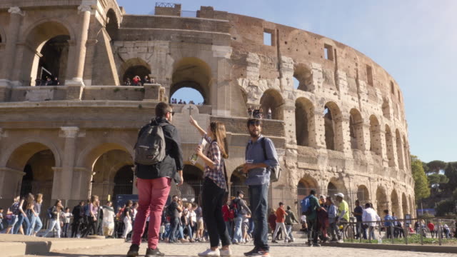 Three young friends tourists standing in front of colosseum in rome reading map guide for directions pointing with backpacks sunglasses happy beautiful girl long hair