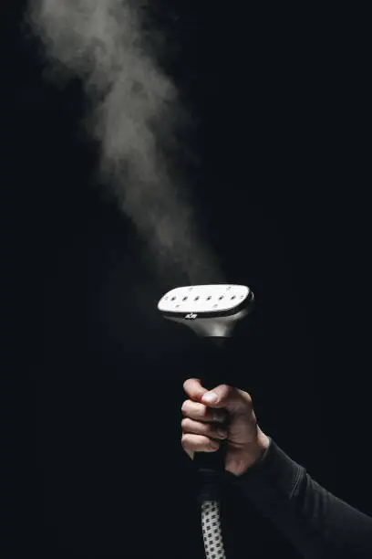 Photo of close-up partial view of person holding garment steamer with steam on black