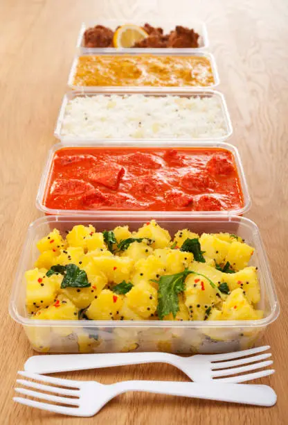 Takeaway Indian Food  - a selection of Indian takeaway food in plastic containers on a wooden table. Aloo saag (potato spinach curry), chicken tikka masala,basmati rice  chicken bhoon or  bhuna, and onion bhaji or pakora.