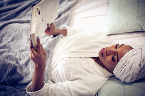 African American woman lying in bed and reading book. Space for copy.