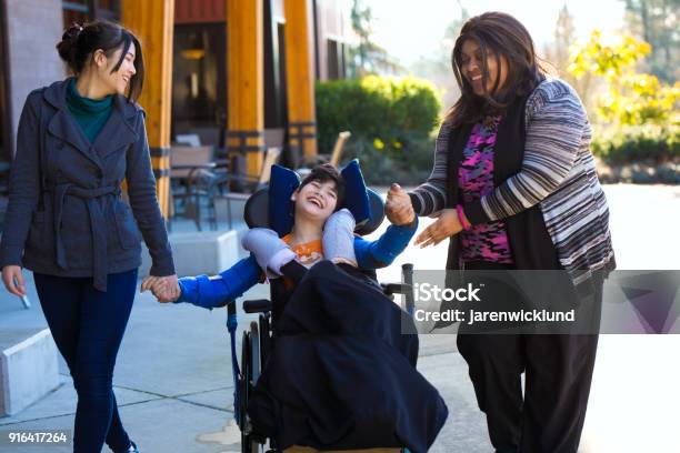 Disabled Boy In Wheelchair Holding Hands With Caregivers On Walk Stock Photo - Download Image Now