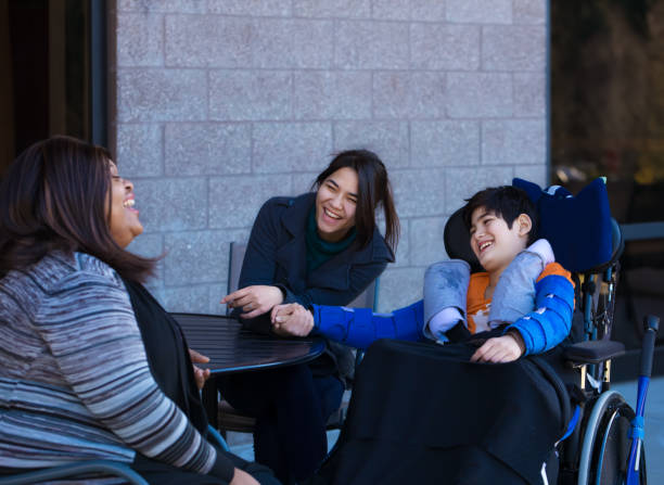 Disabled boy in wheelchair at table outdoors talking with caregivers stock photo