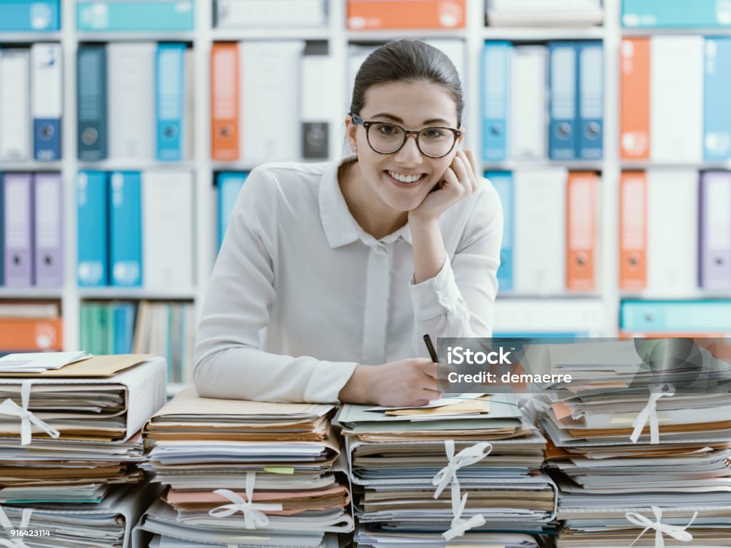 Smiling office clerk and piles of paperwork Smiling young office worker leaning on piles of paperwork File Folder Stock Photo