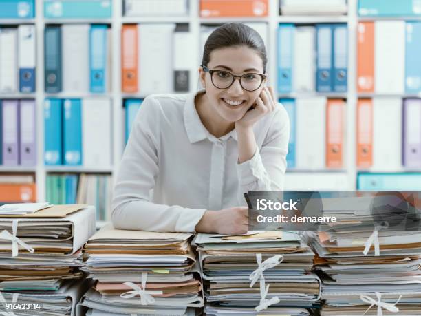 Smiling Office Clerk And Piles Of Paperwork Stock Photo - Download Image Now - File Folder, Desk, Office