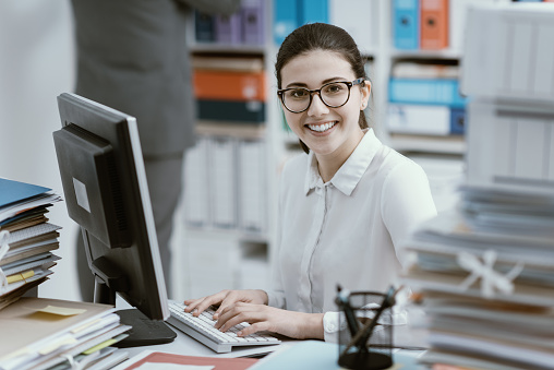 Young smiling secretary working at office desk and stacks of paperwork