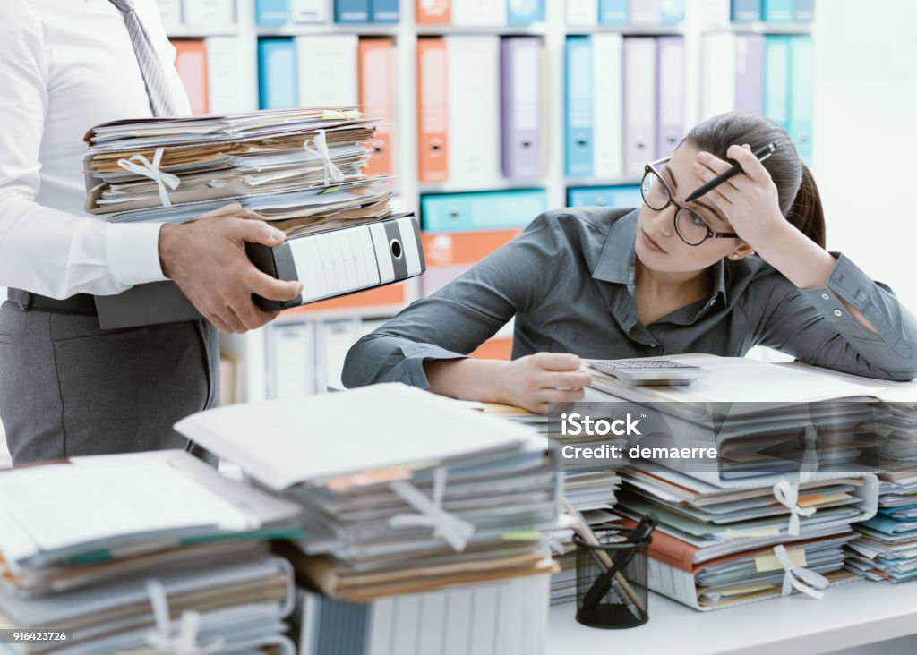 Young secretary overwhelmed by work Young stressed secretary in the office overwhelmed by work and desk full of files, her boss is bringing more paperwork to her Emotional Stress Stock Photo