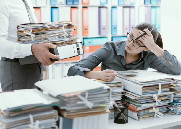 Young secretary overwhelmed by work Young stressed secretary in the office overwhelmed by work and desk full of files, her boss is bringing more paperwork to her managing stress stock pictures, royalty-free photos & images