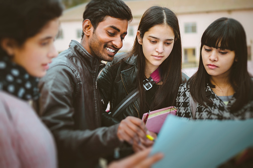 Outdoor image of happy Asian, Indian multi ethnic young adult college students discussing project together on chart paper in college campus at day time. Shoot location Thodo Ground, Solan, (a city in mountain) Himachal Pradesh, India.