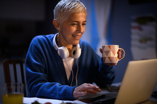 Senior woman working from home late at night. She is having a cup of coffee while working on laptop computer.