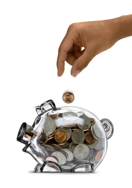 Ethnic female's hand dropping a coin into a clear piggy bank full of coins. Image composed of several shots and composed in Photoshop.