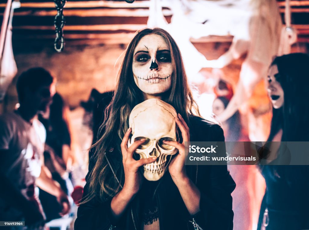 Young woman with skeleton make-up holding skull at Halloween party Young woman with santa-muerte make-up and disguise holding skull at Halloween dungeon party Halloween Stock Photo