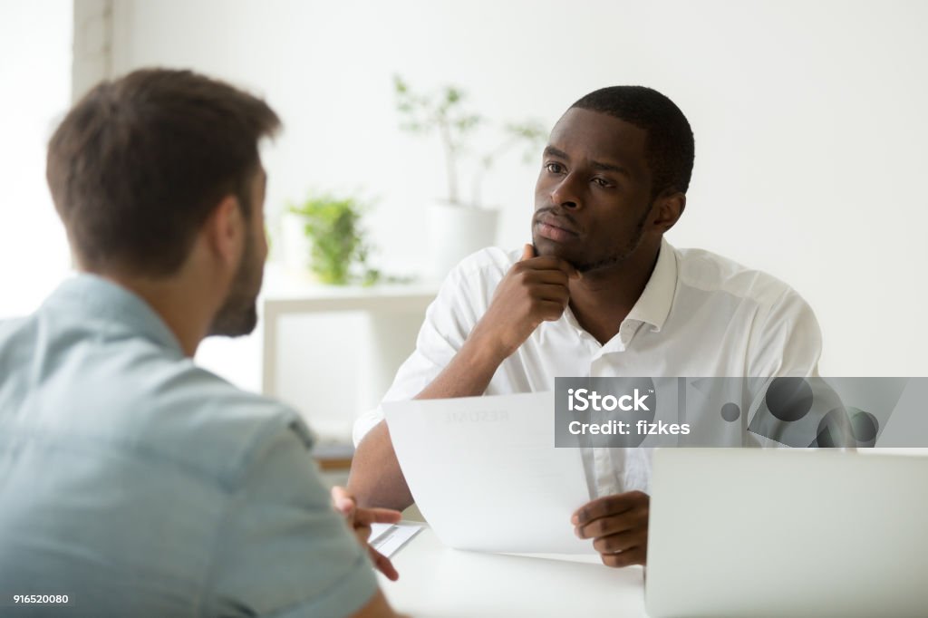 Serious attentive african hr listening to candidate at job interview Serious attentive african hr manager listening to caucasian candidate at job interview, focused strict employer thinking having doubt about hiring decision while talking to male applicant in office Listening Stock Photo