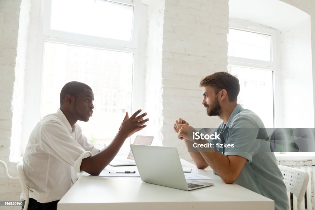 African-american and caucasian colleagues brainstorming new startup idea at work Multiracial businessmen negotiate discuss project talking at work, african and caucasian colleagues brainstorm new idea in office, black entrepreneur explaining corporate teamwork to white coworker Discussion Stock Photo