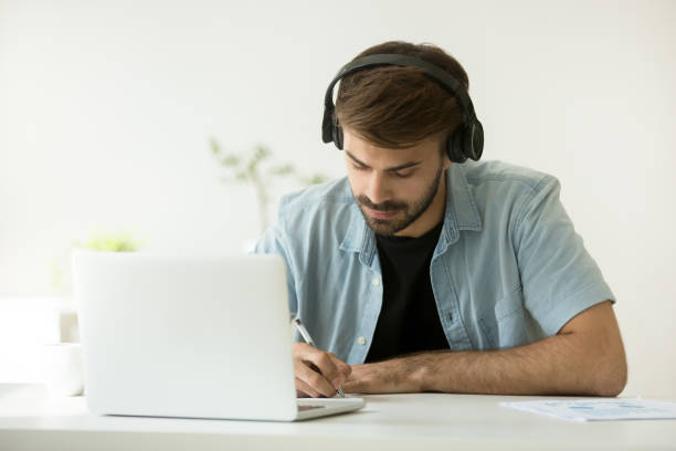 istock Focused man wearing headphones writing notes learning online with laptop 916520196